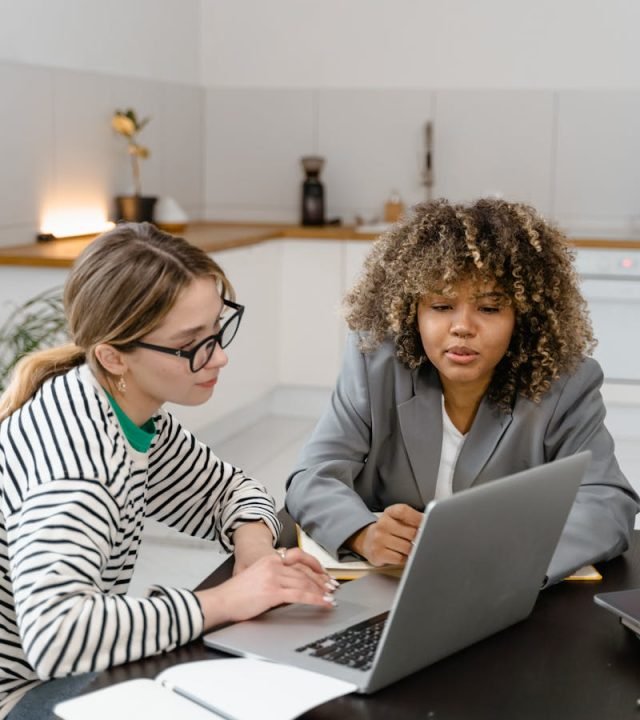 Two women discuss work strategy at a laptop in a modern office.