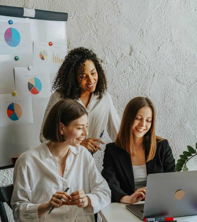 Three businesswomen collaborate with graphs and a laptop in a modern office setting.